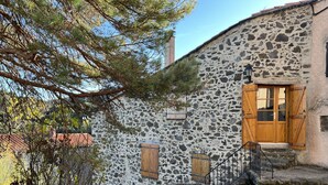 Ein rustikales Steinhaus mit Holztür und Fensterläden, einer Steintreppe und einem Baum im Vordergrund.