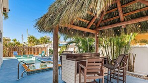 A pool area with a wooden bar and chairs, surrounded by palm trees and a thatched roof structure. - LJ's Cottages 3 - BALCONY BEACH POOL SAND SUNSETS (Sarasota)