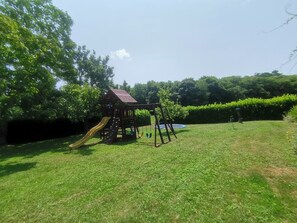 Una estructura de juego de madera con tobogán y pared de escalar, rodeada de un césped cuidado y árboles frondosos.