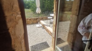A view through a glass door of an outdoor area with a stone wall, gravel ground, and a white umbrella. - Gite Tout Confort 4personnes Avec Piscine, Proche Lot, Dordogne, Lot et Garonne (Marminiac)