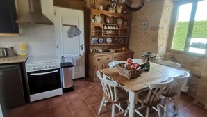 A rustic kitchen with a stove, oven, and hanging pots. There is a wooden table with chairs and a basket of snacks. - Gite Tout Confort 4personnes Avec Piscine, Proche Lot, Dordogne, Lot et Garonne (Marminiac)