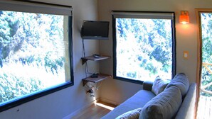 Ein gemütliches Wohnzimmer mit einer Couch, einem Holztisch und einem Fernseher. Große Fenster sorgen für natürliches Licht und Blick auf Bäume.