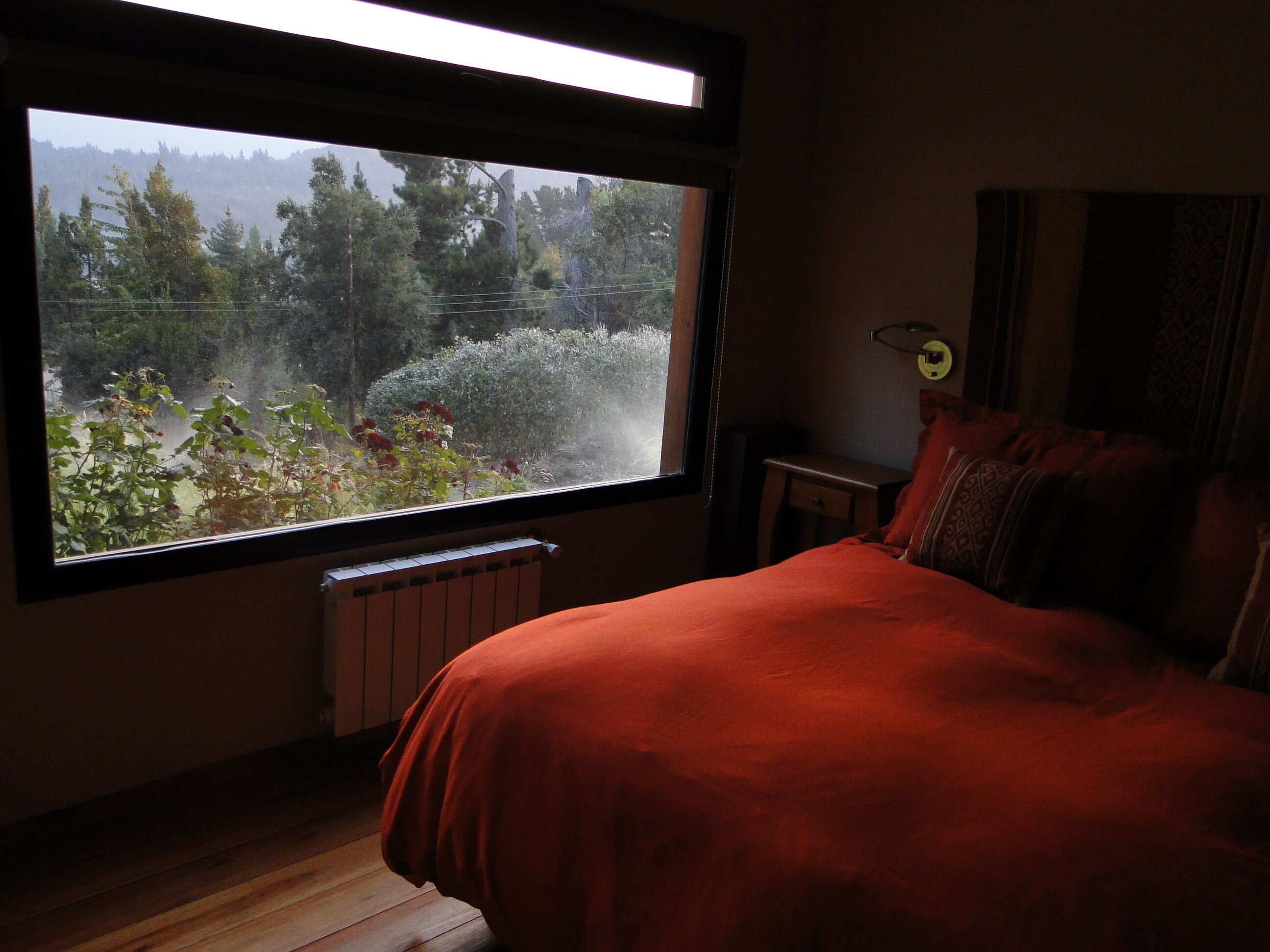 A bedroom with a large window, a bed with a red blanket, a radiator, and a small bedside table.