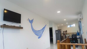 A modern living room with a large flat-screen TV mounted on the wall, a wooden shelf with decorative items, and a unique blue whale mural. - My place in paradise (Puerto Baquerizo Moreno)