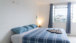 A bedroom with a bed, pillows, a bedside table with a lamp, a window with curtains, and a view of buildings outside. - My place in paradise (Puerto Baquerizo Moreno)