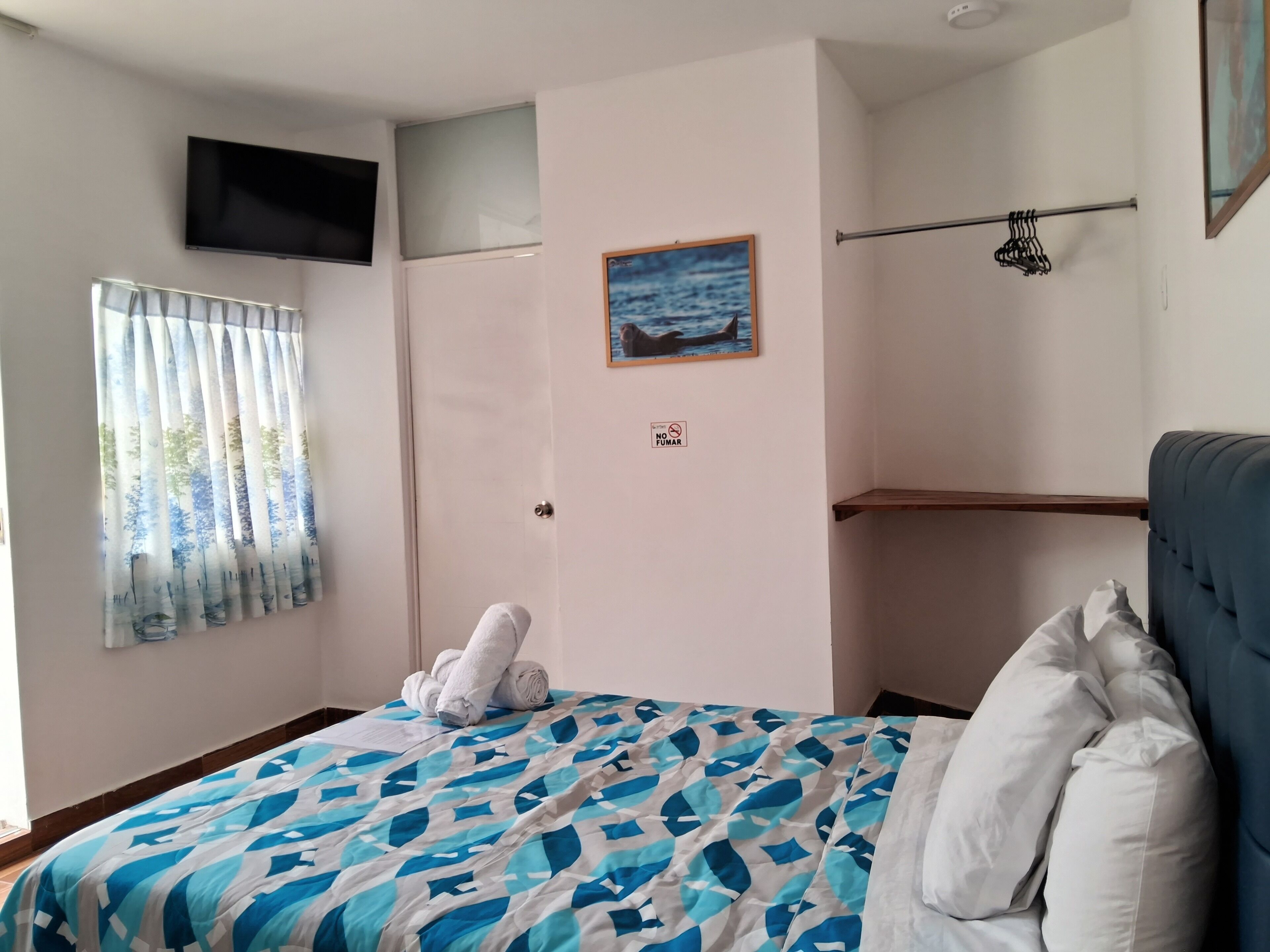 Un dormitorio con cama, televisor montado en la pared, ventana con cortinas, un cuadro y una toalla sobre la cama.