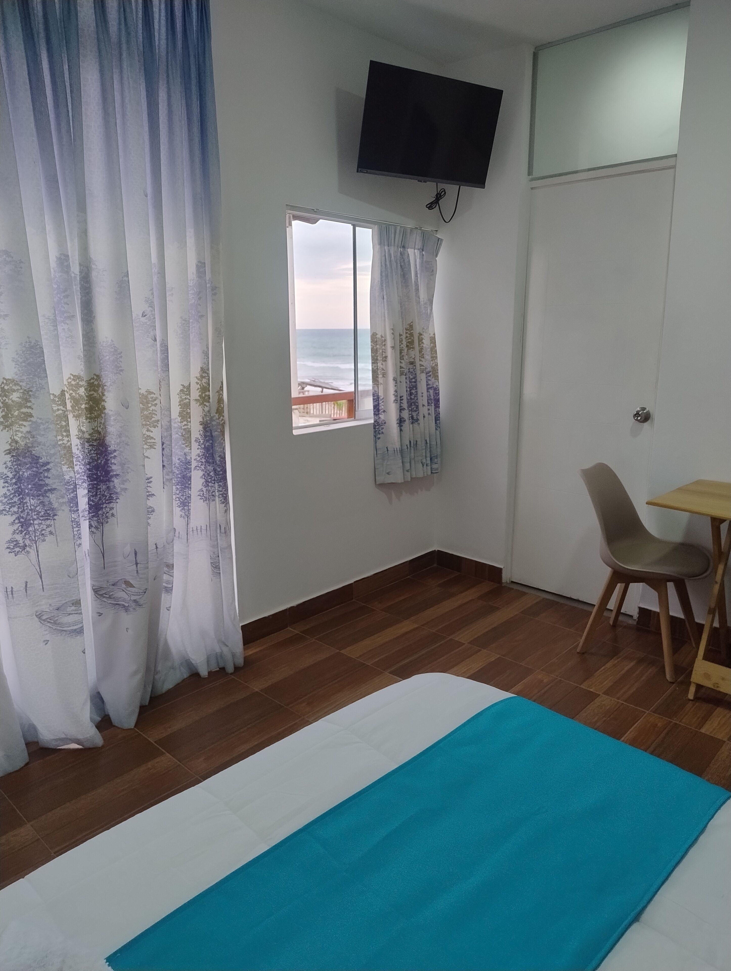 A room with a bed, a TV mounted on the wall, a window with a view of the sea, and a small table with a chair.
