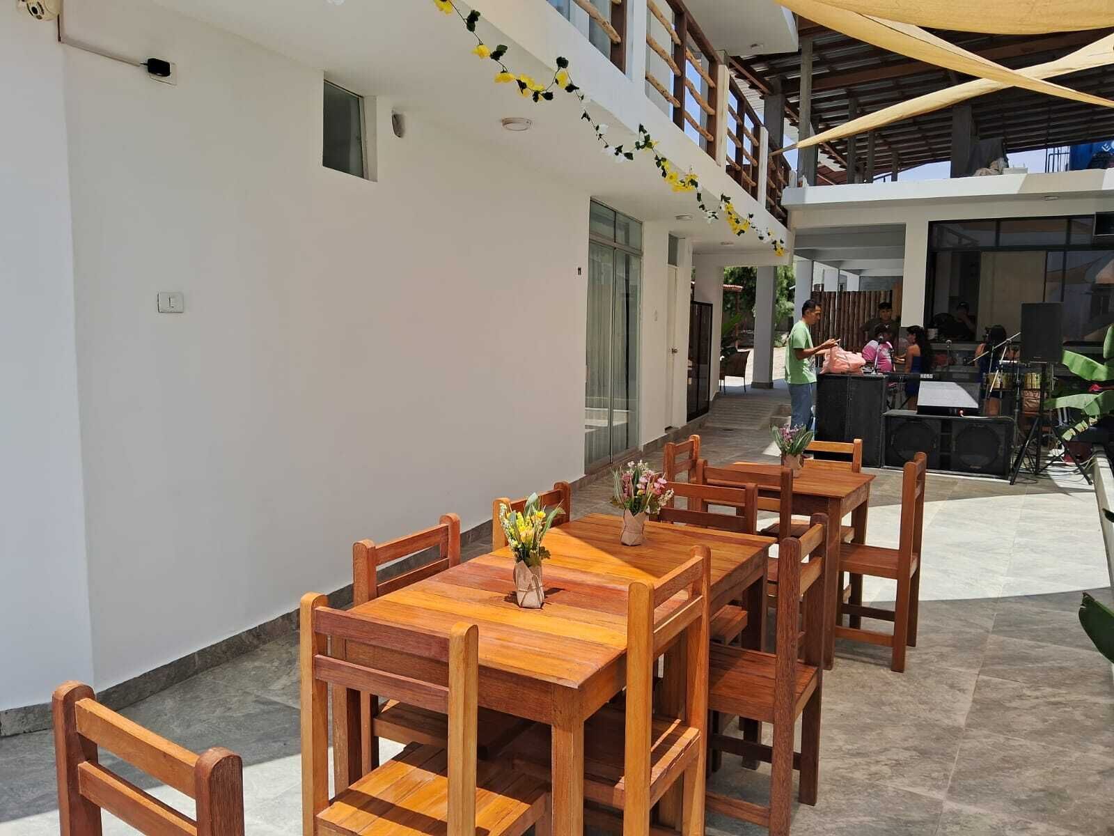 Un área de comedor al aire libre con mesas y sillas de madera, un pequeño arreglo floral en cada mesa, y un espacio de estar techado al fondo.
