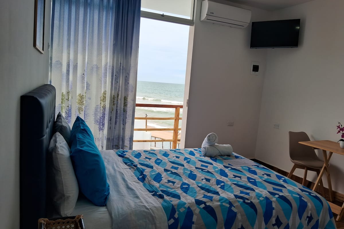 A hotel room with a bed, a TV, a balcony with a view of the sea, and a small table with a plant.