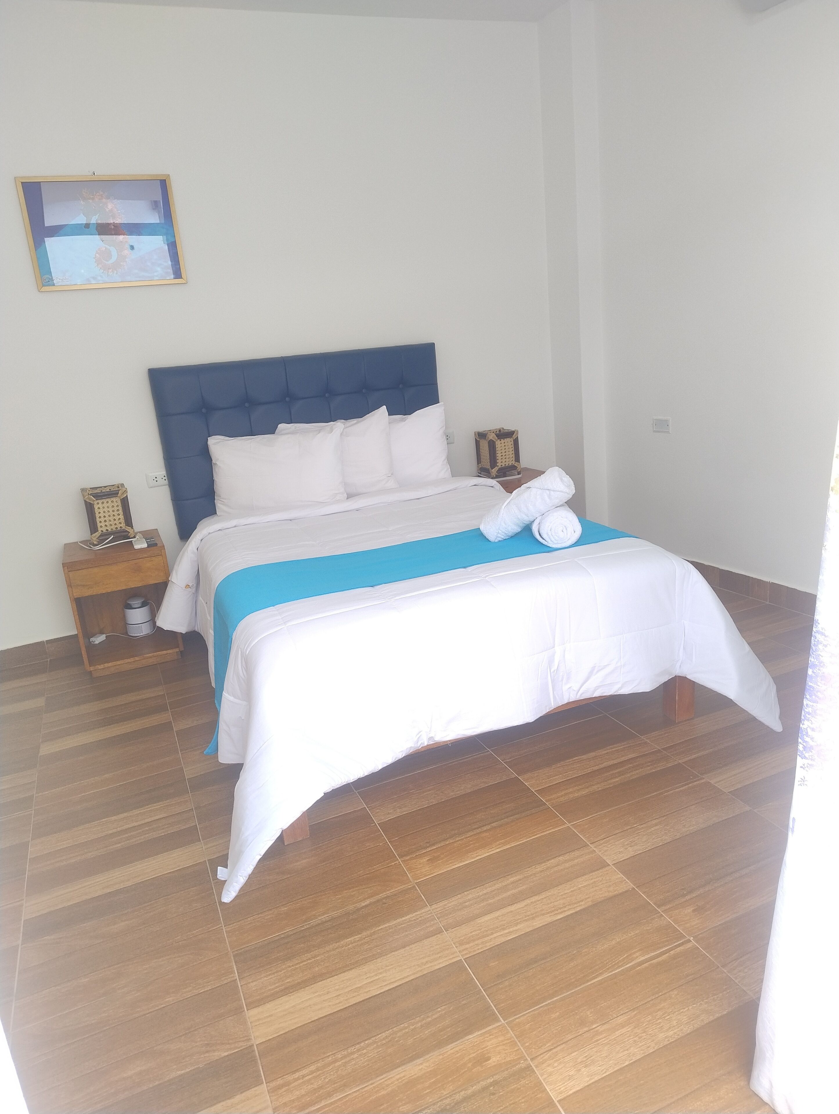 A neatly made bed with a blue headboard, white bedding, and a blue accent on the bedspread. There is a nightstand with a lamp and a small decorative item beside the bed.