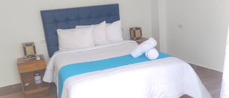 A neatly made bed with a blue headboard, white bedding, and a blue accent on the bedspread. There is a nightstand with a lamp and a small decorative item beside the bed.