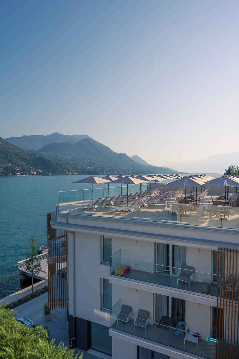 Ein modernes Hotel mit Dachterrasse und Blick auf einen See, ausgestattet mit Liegestühlen und Sonnenschirmen.