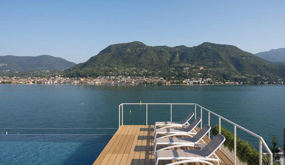 Eine Terrasse mit Liegestühlen mit Blick auf ein Gewässer, vor dem sich ein Gebirge erhebt.