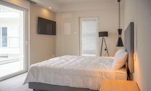 Een slaapkamer met een bed, nachtkastje, lamp, televisie aan de muur en een raam met jaloezieën.