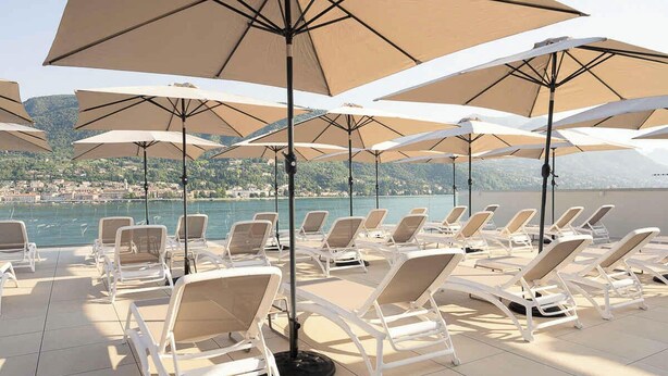 Un espace extérieur lumineux, aménagé avec des fauteuils et des parasols, offrant une vue sur un plan d’eau et une ville située au loin.