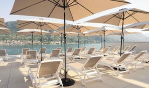 A sunlit outdoor seating area with lounge chairs and umbrellas, overlooking a body of water and a distant town.