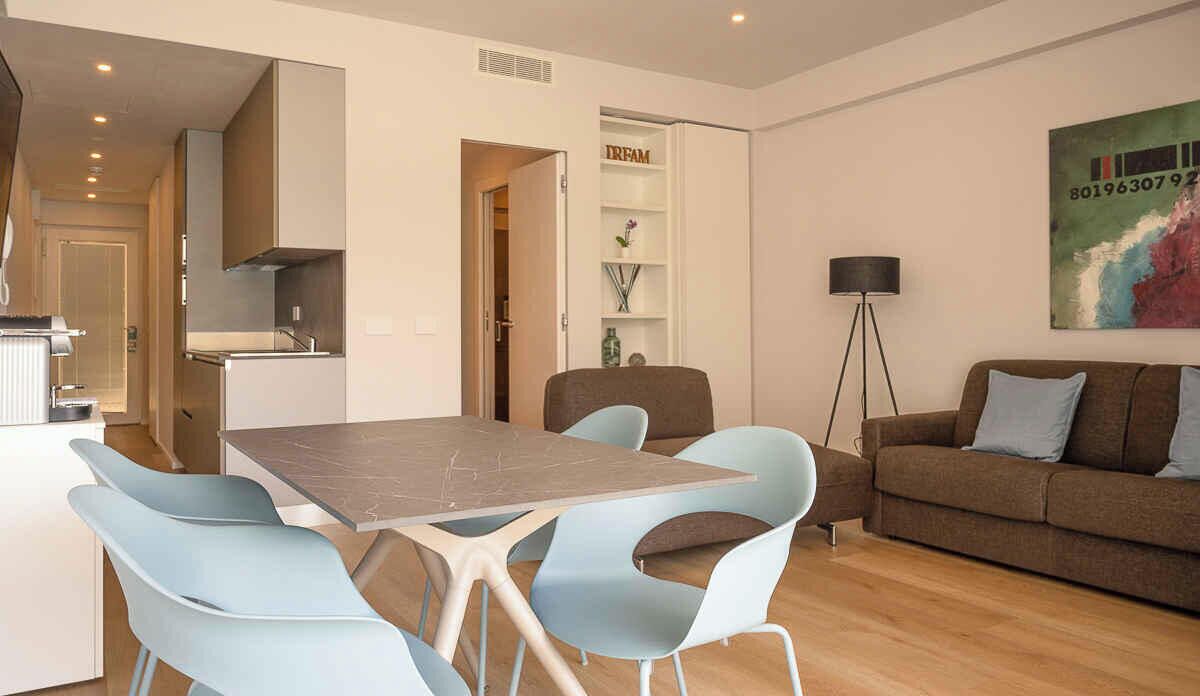 A modern living room with a brown sofa, light blue chairs, a dining table, and a kitchen area with cabinets and a countertop.
