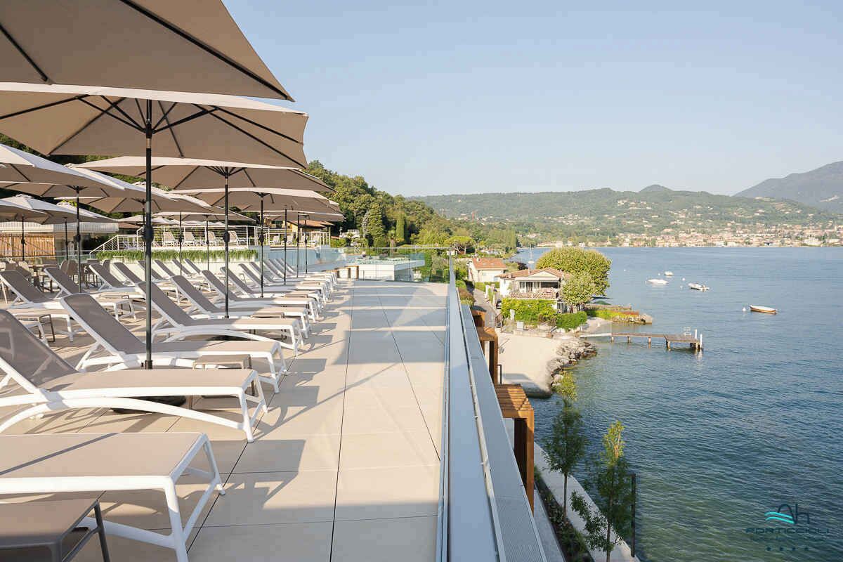 Une terrasse au bord de l’eau, dotée de chaises longues et de parasols, qui offre une vue sur un lac où naviguent des bateaux et sur une ville située au loin.