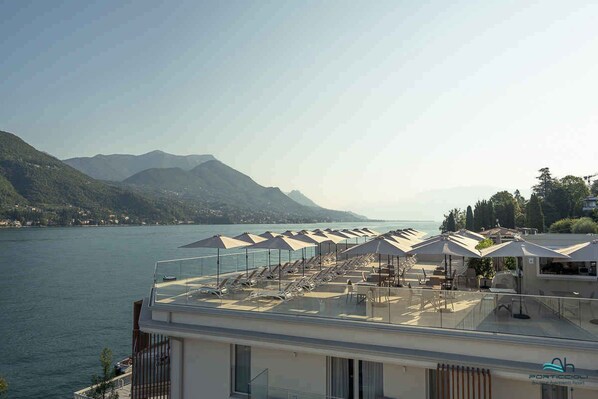 A rooftop terrace with sun loungers and umbrellas overlooking a lake and mountains.