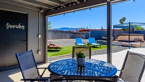 Une terrasse avec une table ronde et des chaises, une petite plante en pot, et une vue sur un jardin avec une piscine, des chaises longues et un jacuzzi.