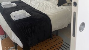 A neatly made bed with white and black bedding, a black throw blanket, and two folded towels. There is a framed picture on the wall above the bed, a bedside lamp, and a window with sheer curtains.