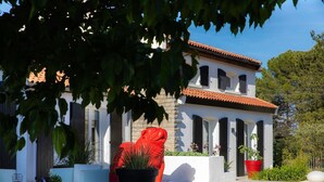 A white building with a red roof, columns, and a statue of a red figure. There are potted plants and a bench in the foreground.