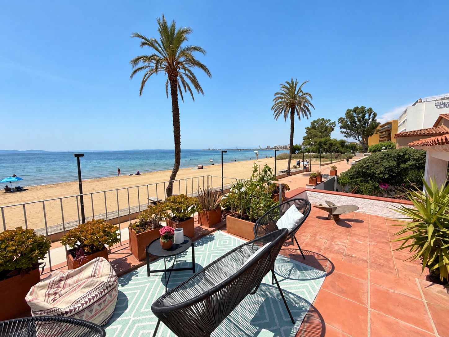 A terrace with outdoor seating, a view of the beach, and palm trees.