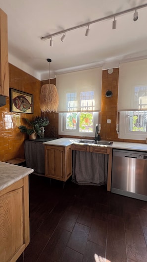 A modern kitchen with wooden cabinetry, a built-in dishwasher, a large window with a view of greenery, and a hanging light fixture.