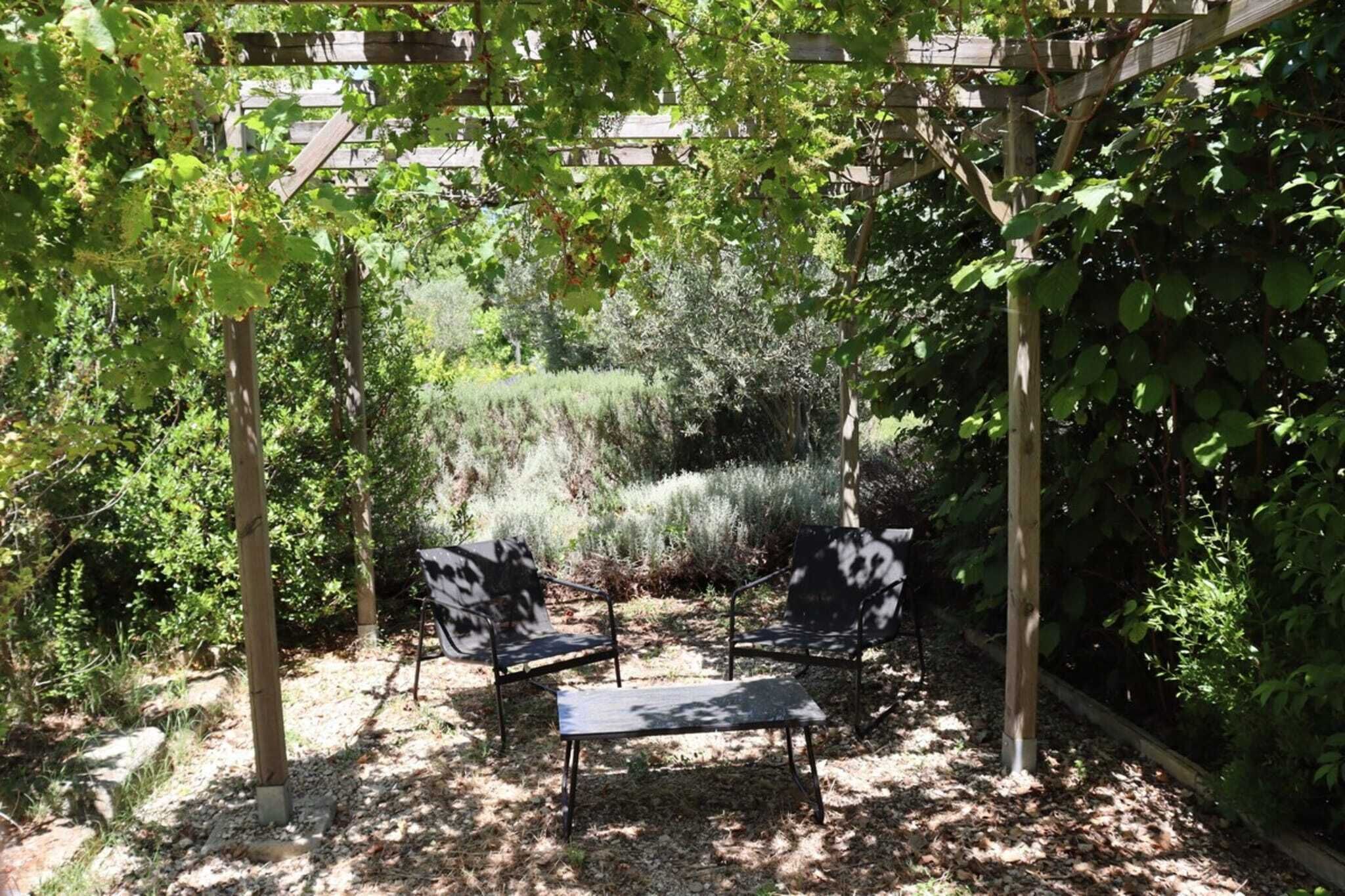 Un espacio de estar al aire libre cubierto con dos sillas y una mesa, rodeado de abundante follaje y una estructura de pérgola de madera.
