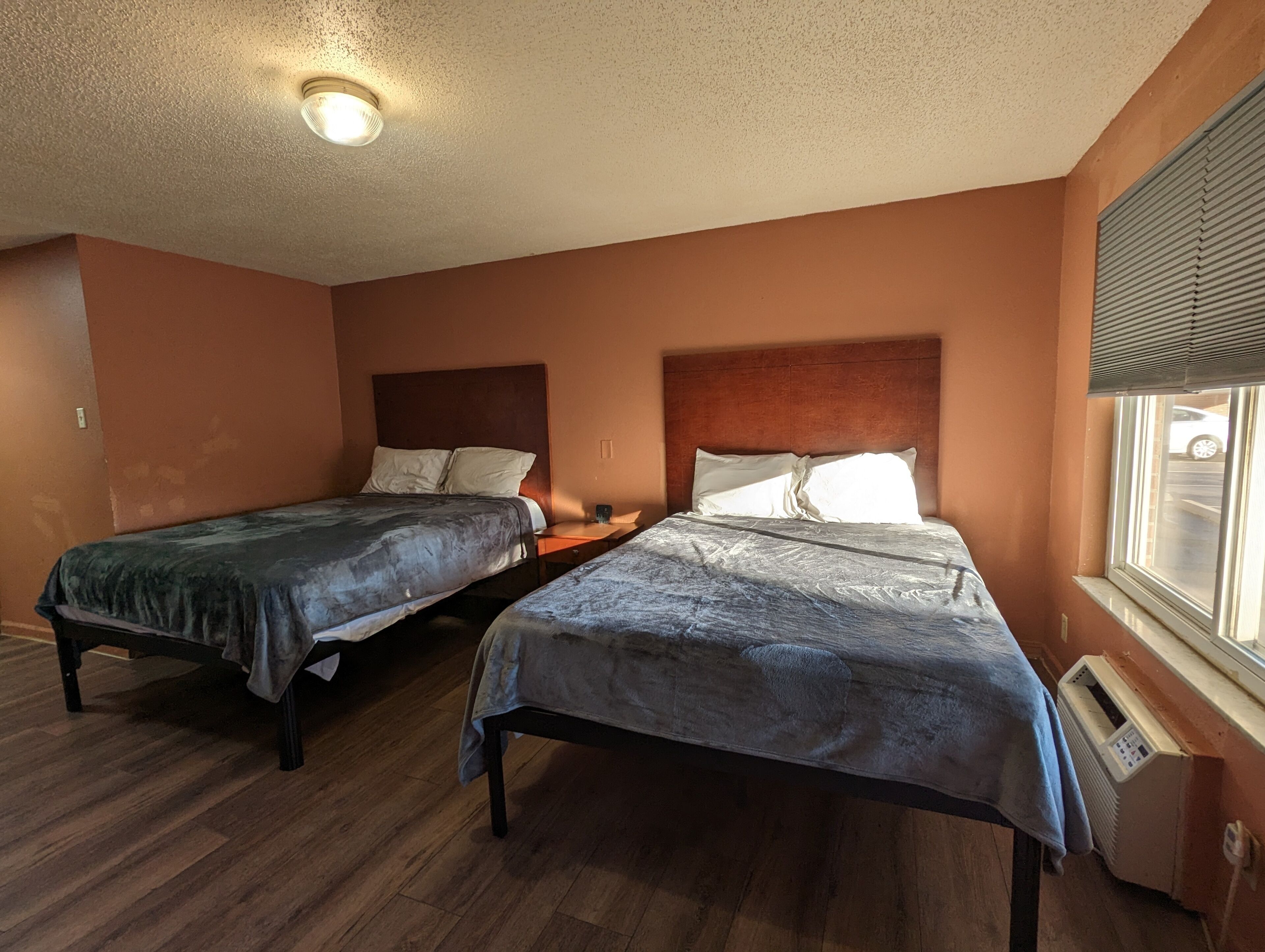 A room with two beds, wooden flooring, a ceiling light, and a window with blinds.