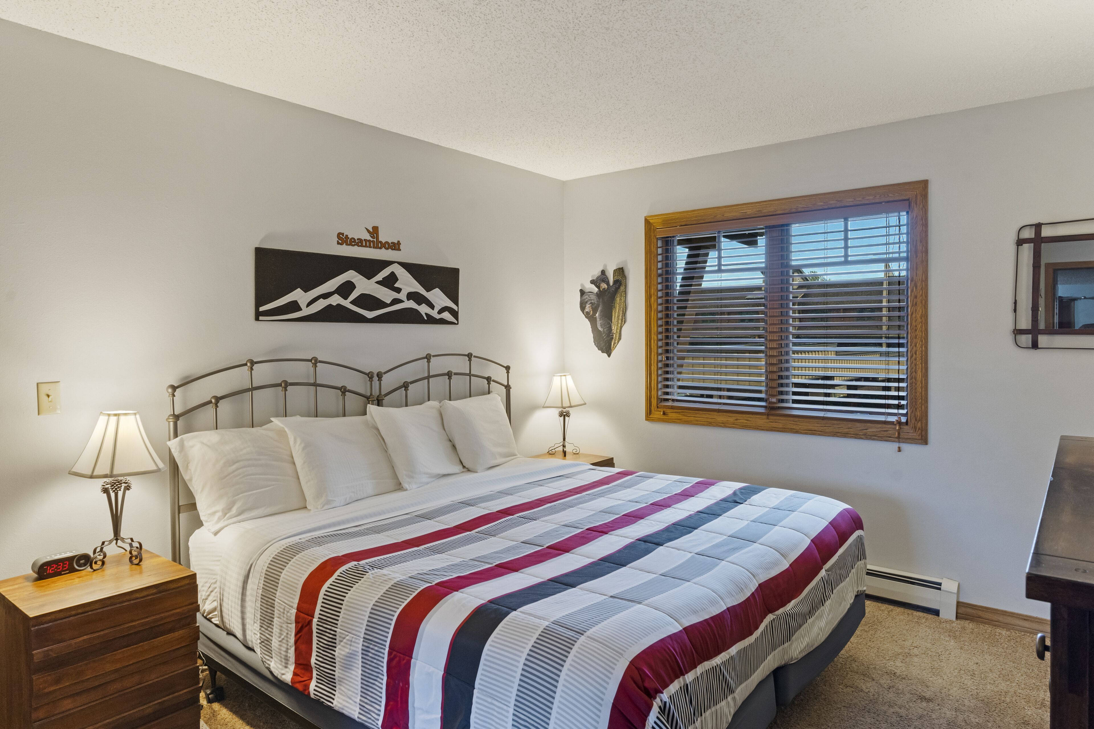 A bedroom with a bed, two bedside tables with lamps, a wall clock, a mountain-themed artwork, a window with blinds, and a mounted picture frame.