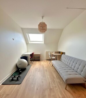 Um apartamento moderno no estilo loft, com teto inclinado, claraboia, sofá cinzento, pequena escrivaninha e equipamentos de ginástica.