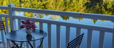 Varanda com mesa e cadeiras, um vaso com flores e vista para um lago rodeado por árvores.