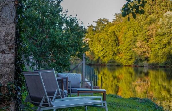Uma área de estar à beira do lago com cadeiras e uma mesa, rodeada por vegetação exuberante e um lago tranquilo.