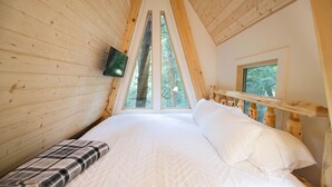 A cozy bedroom with a wooden sloped ceiling, a large bed with white bedding, a TV mounted on the wall, and a window with a view of trees. - Peaceful Pines Luxury Treehouse (Lucinda)