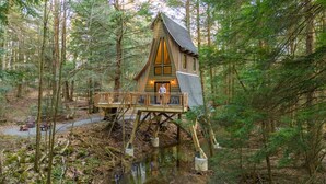 A wooden cabin with a balcony, situated in a forested area with a small stream. - Peaceful Pines Luxury Treehouse (Lucinda)