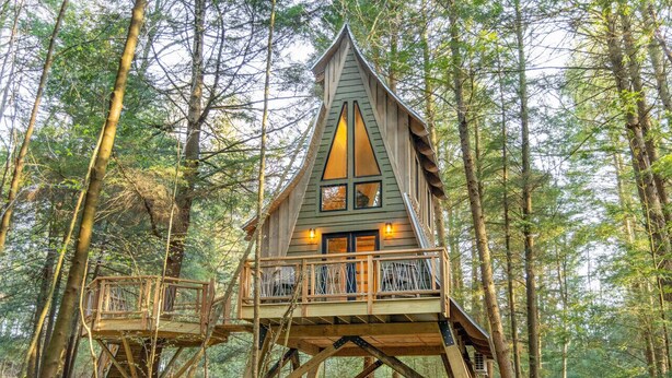 A wooden treehouse with a balcony and a staircase, surrounded by a forest. - Peaceful Pines Luxury Treehouse (Lucinda)