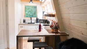 A compact kitchen with wooden walls, a window, and a small dining area. - Peaceful Pines Luxury Treehouse (Lucinda)