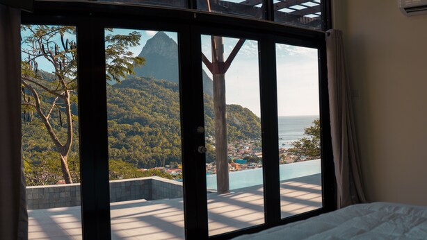 A room with a large window overlooking a mountainous landscape and a pool. - Enclave Villa V3 -Overlooking Pitons & Ocean ! Wow.  (Soufriere)