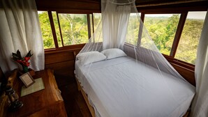 A bedroom with a wooden bed, white bedding, a nightstand with a lamp, and a view of greenery through large windows. - Alta Sanctuary (Tambopata)