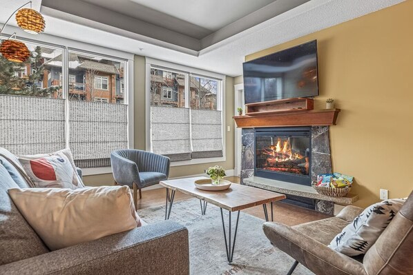 A cozy living room with a fireplace, comfortable seating, and large windows offering a view of the outside.