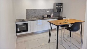 A modern kitchen with a built-in oven, a microwave, and a coffee maker on the countertop. There is a dining table with two chairs. - Bienvenue à la Maison D'hôtes L'instant Present (Le Moule)