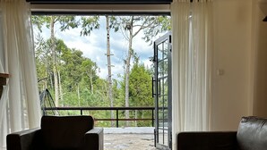 Un salón con sofá, sillón y mesa de centro, una amplia puerta corredera de vidrio que da a un balcón con vistas a árboles.