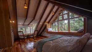Un dormitorio con suelo de madera, una cama con ropa de cama blanca, un asiento junto a la ventana con un cojín y una silla.