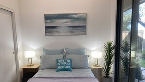 Une chambre à coucher avec un lit, deux tables de chevet avec des lampes, un tableau au-dessus du lit et une vue sur l’extérieur par une porte vitrée.