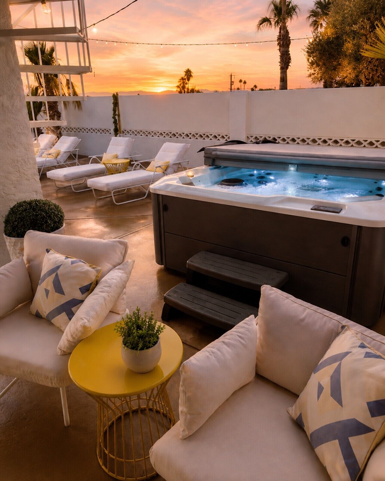 A hot tub with lounge chairs and a small table with a plant, set against a sunset backdrop.