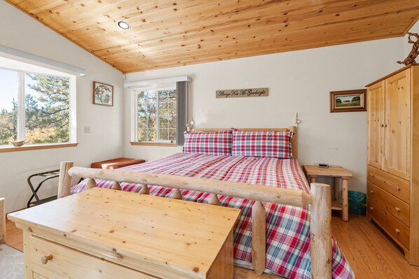 A bedroom with a wooden bed frame, plaid bedding, a wooden dresser, and a view of trees through large windows. - Luxurious Hyes in the Woods (Big Bear City)