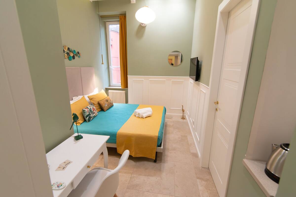 Un dormitorio con cama, mesita de noche y una silla. Se ve una ventana con cortinas, una puerta y un televisor en la pared.