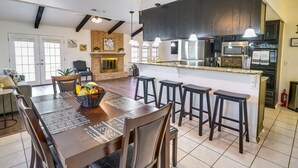 A modern kitchen with dark cabinetry, a central island with stools, a dining area with a long table and chairs, and a fireplace.