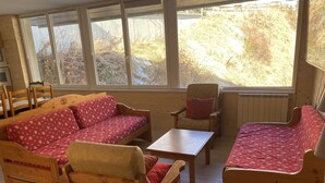 A cozy room with wooden furniture, including a sofa, chairs, and a table. Large windows provide a view of the outdoors. - L'écrin, Appartement aux Pieds des Pistes (Orcières)